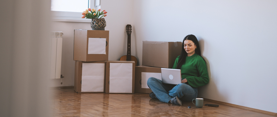 Woman sitting on floor beside a pile of moving boxes using a laptop.
